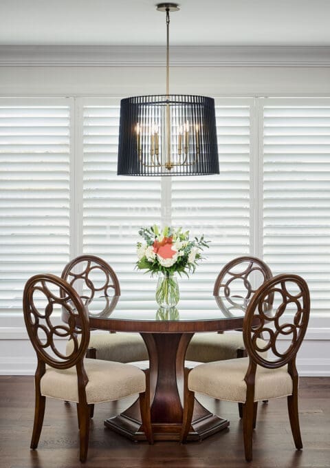 Round wood dining table with sculptural chairs and a statement black pendant fixture, framed by white shutters and soft natural light.