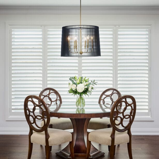 Round wood dining table with sculptural chairs and a statement black pendant fixture, framed by white shutters and soft natural light.