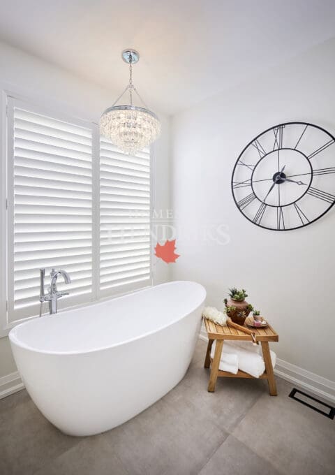 Elegant ensuite with a freestanding oval tub beneath a crystal chandelier, accented by a black roman numeral wall clock and wooden stool with spa décor.