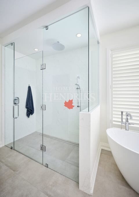 Modern glass-enclosed shower featuring rainfall and handheld fixtures, white marble-look walls, and natural light from plantation-shuttered windows.