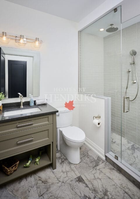 Compact guest bath with olive vanity, black quartz countertop, frameless glass shower, and marble-patterned tile flooring.