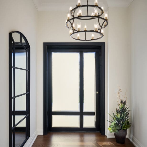 Front Foyer Bright entryway with black-framed glass doors, two-tier chandelier, large arched mirror, and warm wood flooring.