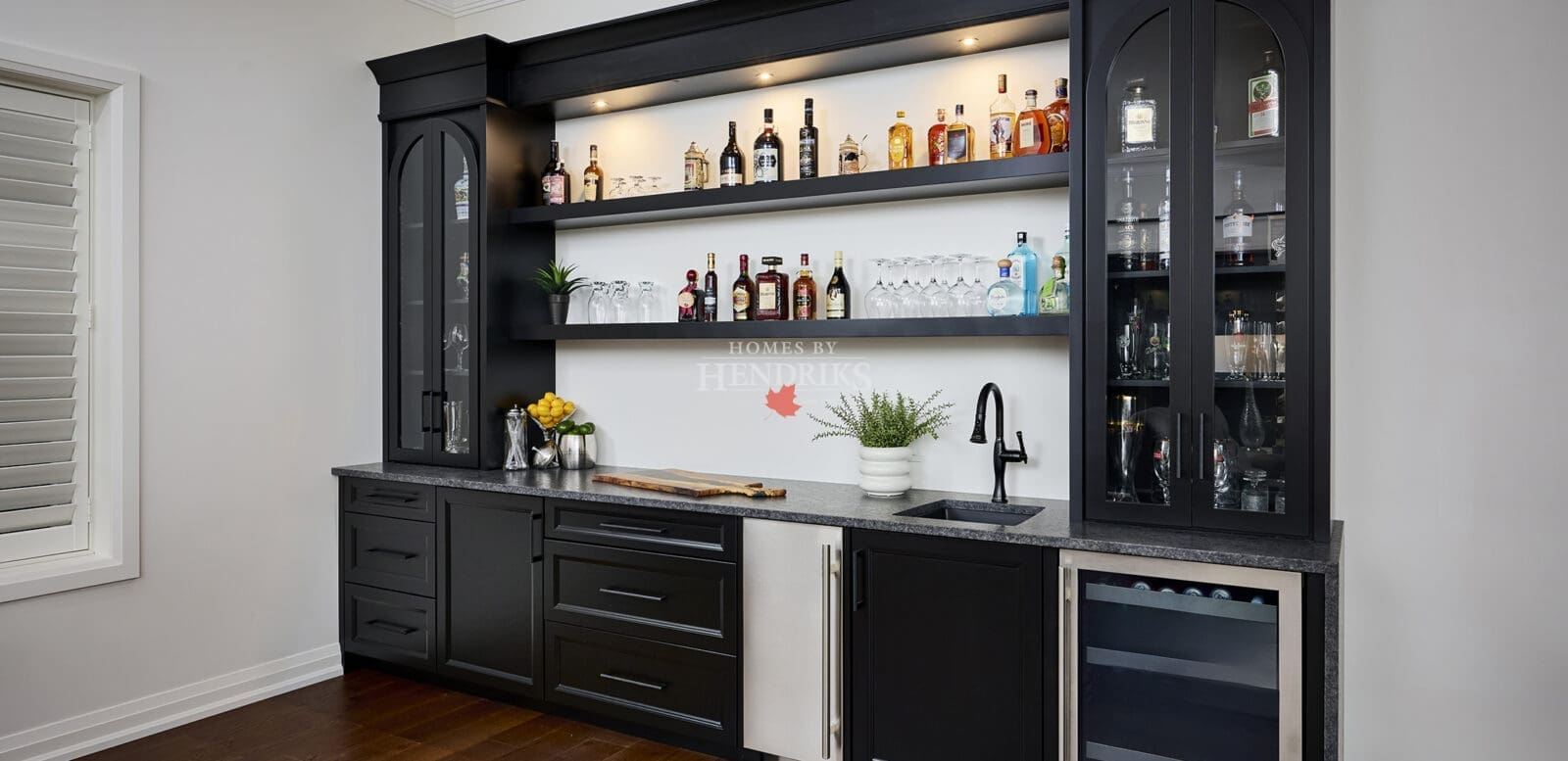 Sophisticated black built-in home bar with glass shelving, beverage fridge, and underlit display of spirits, designed by Homes By Hendriks.
