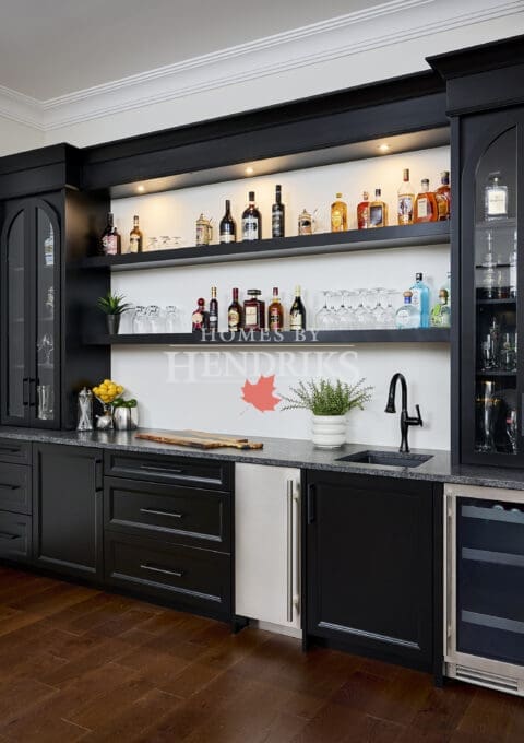 Sophisticated black built-in home bar with glass shelving, beverage fridge, and underlit display of spirits, designed by Homes By Hendriks.