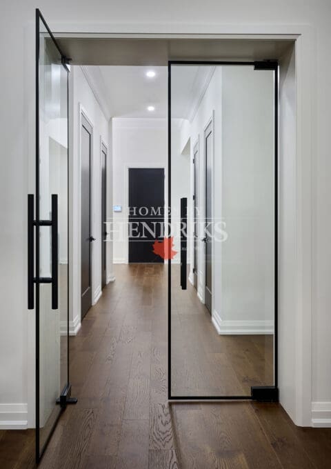 Modern hallway framed by full-height glass doors with black trim and hardware, opening to a minimalist corridor with white walls and oak floors.