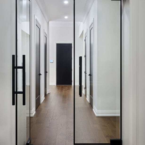 Modern hallway framed by full-height glass doors with black trim and hardware, opening to a minimalist corridor with white walls and oak floors.